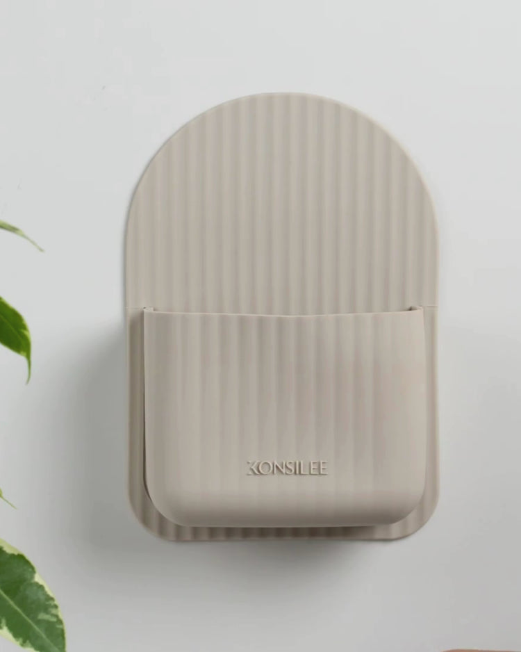 A hand using a damp, dark-purple microfiber cloth to wipe the surface of a soft-brown, arch-shaped silicone shower caddy. The close-up highlights the vertical fluted texture and an embossed 'Konsilee' logo on the front, demonstrating the easy-to-clean material. The wall is a solid off-white shade, with a green vine plant visible to the side.